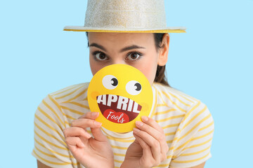 Young woman holding paper smile with text APRIL FOOLS on blue background
