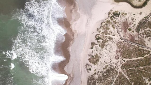 DRONE AERIAL FOOTAGE: Northern Littoral Natural Park in Esposende, Portugal. Sea, beach boulders, pebble shore, and waves. Sea waves breaking on rocks. Sunny beach with sand dunes and a cloudy sky.