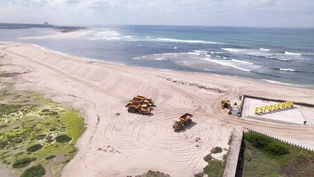 Esposende, Portugal, April 10, 2022: DRONE AERIAL FOOTAGE - The two sides of Restinga de Ofir. Esposende fishing harbor maintenance dredging. Dumper vehicles and rotating excavator parked on the sand.