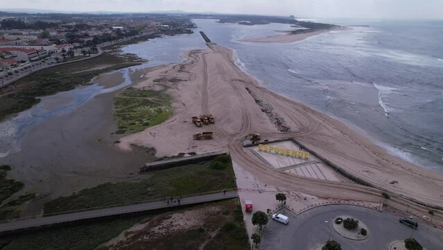 Esposende, Portugal, April 10, 2022: DRONE AERIAL FOOTAGE - The two sides of Restinga de Ofir. Esposende fishing harbor maintenance dredging. Dumper vehicles and rotating excavator parked on the sand.