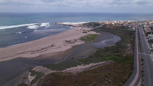 Esposende, Portugal, April 10, 2022: DRONE AERIAL FOOTAGE - The two sides of Restinga de Ofir. Esposende fishing harbor maintenance dredging. Dumper vehicles and rotating excavator parked on the sand.