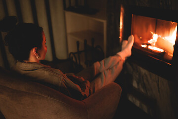 Cozy night in the cabin by the fireplace, fireplace burns in the scandinavian cottage chalet house, burning fire with charcoal and firewood, young woman with a drink reads book