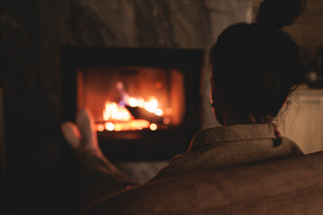 Cozy night in the cabin by the fireplace, fireplace burns in the scandinavian cottage chalet house, burning fire with charcoal and firewood, young woman with a drink reads book