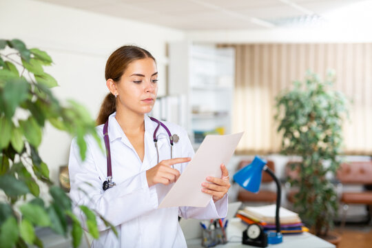 Young Woman Doctor Working In The Clinic Is Attentive Studying The Patient's Outpatient Card, Standing In The Resident's ..office. Close-up Portrait..