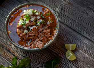 A bowl of BIRRIA or BARBACOA, a traditional Mexican dish with lemons and cilantro on the side over...