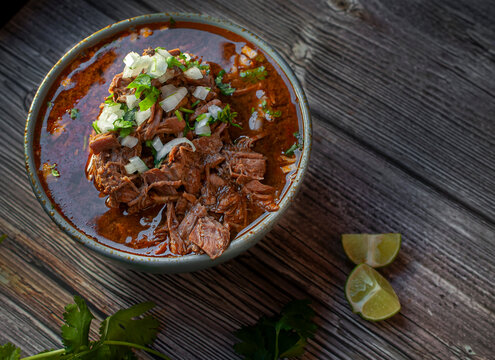 A bowl of BIRRIA or BARBACOA, a traditional Mexican dish with lemons and cilantro on the side over a wooden table, horizontal shot

