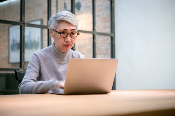 Mature asian business woman wears glasses using laptop computer sit at workplace desk. Happy senior older employee 50s businesswoman. - obrazy, fototapety, plakaty
