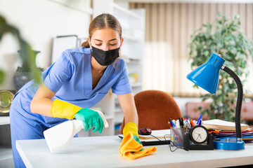 Portrait of a young woman worker of a cleaning company in a protective mask, washing an office desk...