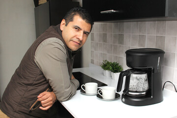 Dark-haired Latino adult man prepares a cup of coffee in a coffee maker to smell and taste at breakfast in the kitchen is thoughtful, worried, thoughtful, depressed
