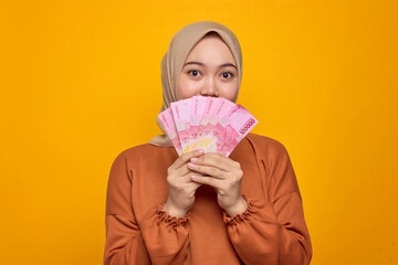 Shocked young Asian woman in orange shirt holding money banknotes isolated over yellow background