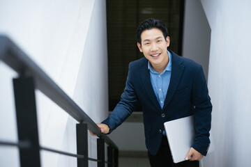 Fototapeta premium Portrait Asian businessman smiling confidently in office stairs with holding laptop.