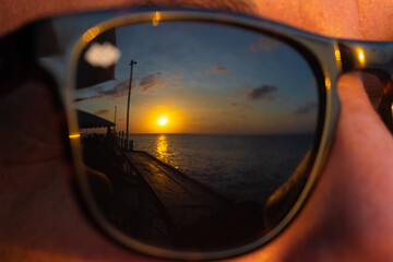 A close up shot of a tropical Caribbean sun setting over the ocean seen through the reflection of a pair of sunglasses on a Caucasian lady's face