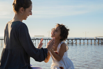 Mother and mixed race adopted daughter playing hand-clapping game together at river pier. Sunset...