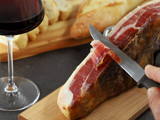 male hand with knife cuts original Jamon Serrano ham into slices, close up, a typical Spanish...