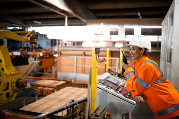 Female worker in safety equipment and hardhat controlling parts assembling in factory. Industrial machines working in background.