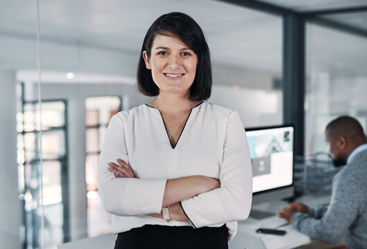 Any Business Challenge You Set, Well Conquer. Cropped Shot Of An Attractive Young Businesswoman Standing With Her Arms Folded While A Colleague Works Behind Her.