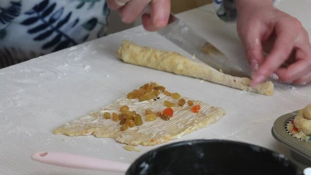 A woman cuts a roll of rolled dough. Prepares cruffin with raisins and candied fruit. Tools and ingredients are laid out on the table..