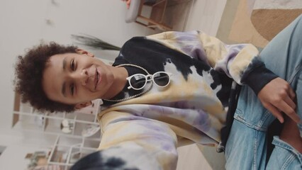 Handheld vertical POV slowmo of Biracial gen z boy in trendy clothes talking and filming himself sitting on rug at his bedroom - Powered by Adobe