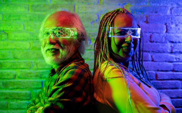 Happy elderly friends having fun with futuristic augmented reality glasses, multiracial couple Asian male and Hispanic American female - Metaverse Concept