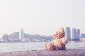 A brown fluffy teddy bear sits on a single bridge looking out to the sea with a copy space background. Lonely, loneliness is pointless.