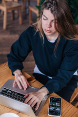 A young beautiful girl in a black jumper in a cafe works with a laptop and a phone