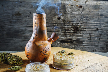 Old brown steamung bong and open cannabis grinder with chopped marijuana weed and medical cannabis in smoke by wooden desck and sackcloth. Horizontal