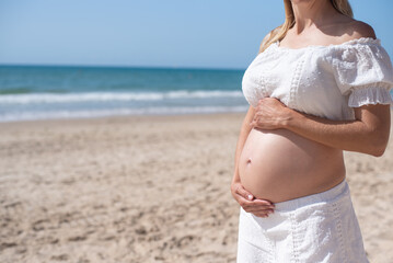 Fototapeta na wymiar Young pregnant woman in white clothes stand on beach and holds hands on belly. Wonderful touching period of waiting baby. Vacation and holiday on sea.