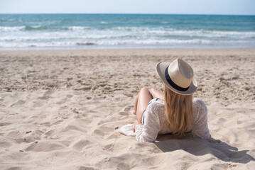 Young woman in white shirt and hat lies on back on sand beach and look far away. Person enjoys summer vacation and holiday on sea.