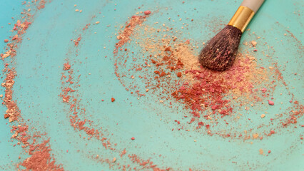 A large blush applicator brush, with a pile of red and cream color makeup powder center around the brush head. On a bluish green background. 