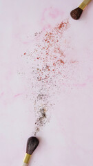 Top view of two makeup brushes, with cosmetic powder spreading out from the brush tip. On a pink marble like surface.
