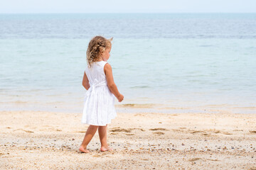 A little girl with curly hair in a white dress walks along a sandy beach on the shore of the sea, ocean and smiles. Sea holidays, travel and beach holidays with children