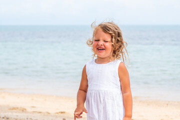 Fototapeta na wymiar A little girl with curly hair in a white dress walks along a sandy beach on the shore of the sea, ocean and smiles. Sea holidays, travel and beach holidays with children