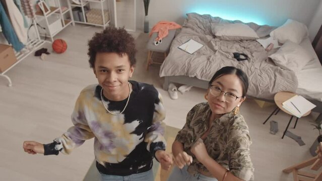 From above slowmo of two cool diverse zoomers looking at camera while having fun and dancing at messy teenage bedroom