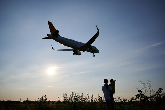 Silhouette of woman hoding kid and looking to landing commercial airplane in the evening. Lifestyle and travel concept.