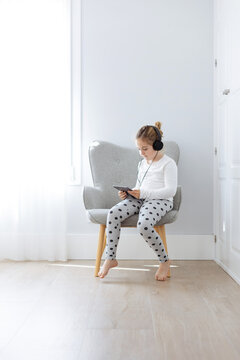 Full body of barefoot girl sitting in armchair and listening to songs from tablet in headphones
