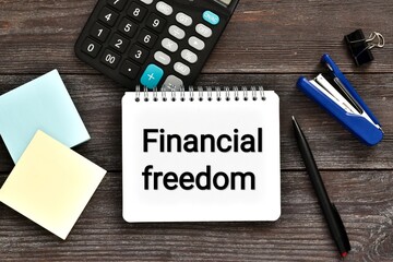 Text FINANCIAL FREEDOM written in notebook with calculator, paper, pen and stapler on wooden table....