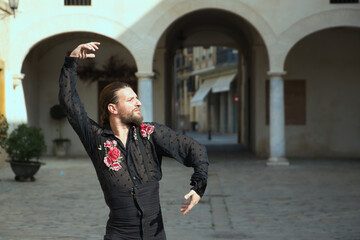 Young man with beard and ponytail, wearing black transparent shirt with black polka dots and red roses, black pants and jacket, dancing flamenco in the city. Concept art, dance, culture, tradition.