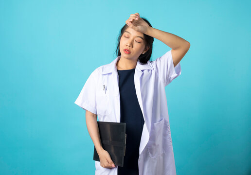 Exhausted young asian female doctor looking tired after shift in hospital, looking sad with fatigue, standing on blue background.