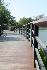 Brown wood walkway and black painted metal fence beside pond in park, Thailand.