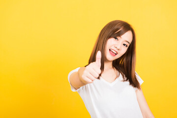 Asian happy portrait beautiful cute young woman teen standing wear t-shirt showing gesturing finger thumb up looking to camera isolated, studio shot on yellow background with copy space