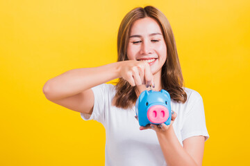 Portrait Asian Thai beautiful happy young woman smiling save money by dropping coin to a piggy bank and looking to piggybank, studio shot isolated on yellow background, with copy space