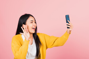 Happy Asian portrait beautiful cute young woman smiling excited  making selfie photo, video call on smartphone studio shot isolated on pink background, female hold mobile phone raise hand say hello