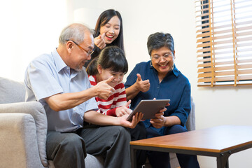 Obraz premium Smiling Asian grandparents on couch with granddaughter looking at tablet. happy three generation family spending time together at home.