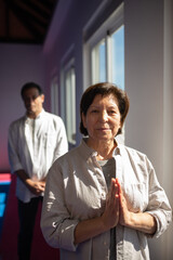 Fototapeta na wymiar Portrait of content Asian yoga coach in gym. Senior woman in shirt holding hands in Namaste. Meditation concept