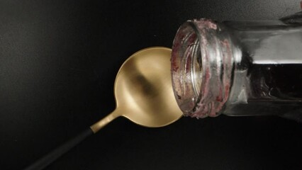 I pour red syrup from a bottle on a golden spoon, black background, top view. Dolly slider extreme close-up. Laowa Probe