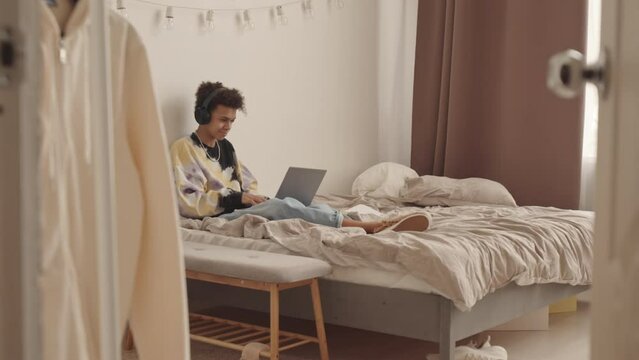 Slowmo Of Biracial 14 Year Old Zoomer Boy Smiling While Typing Messages To His Friends Communicating Online On Laptop Sitting On Bed In His Bedroom