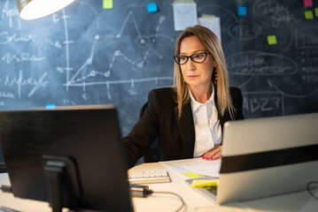 Portrait of businesswoman in workflow. Serious woman working with two computers in evening analyzing financial data, calculating profit for investments. Business growth and investment activity concept