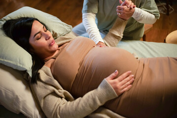 Cropped image of pregnant woman and helpful midwife at home. Woman in casual clothes lying on bed, midwife holding hand. Pregnancy, medicine, home birth concept - obrazy, fototapety, plakaty