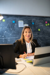 Portrait of businesswoman working in evening. Happy woman working at table in office, making financial analysis writing down results with pen and looking at camera. Finance and business growth concept