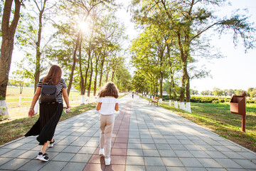 Back view of family walking on trees alley path in the park on sunny autumn day. Mother with her daughter enjoy day in nature.